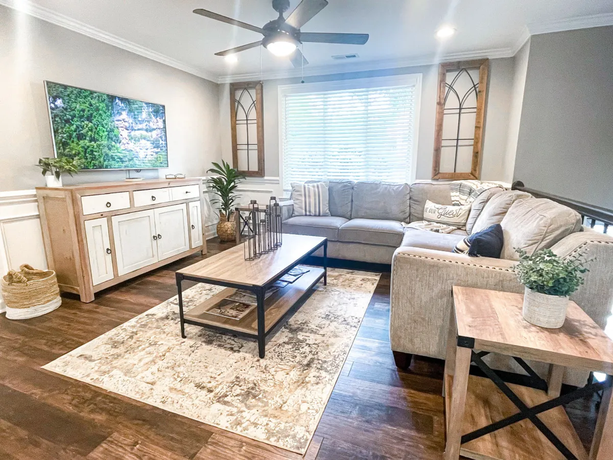 A stylish, sunlit living room featuring a plush sectional sofa, modern coffee table, decorative plants, and a large television. The inviting space is designed for relaxation, family gatherings, and entertainment, with warm decor and comfortable seating ideal for groups.