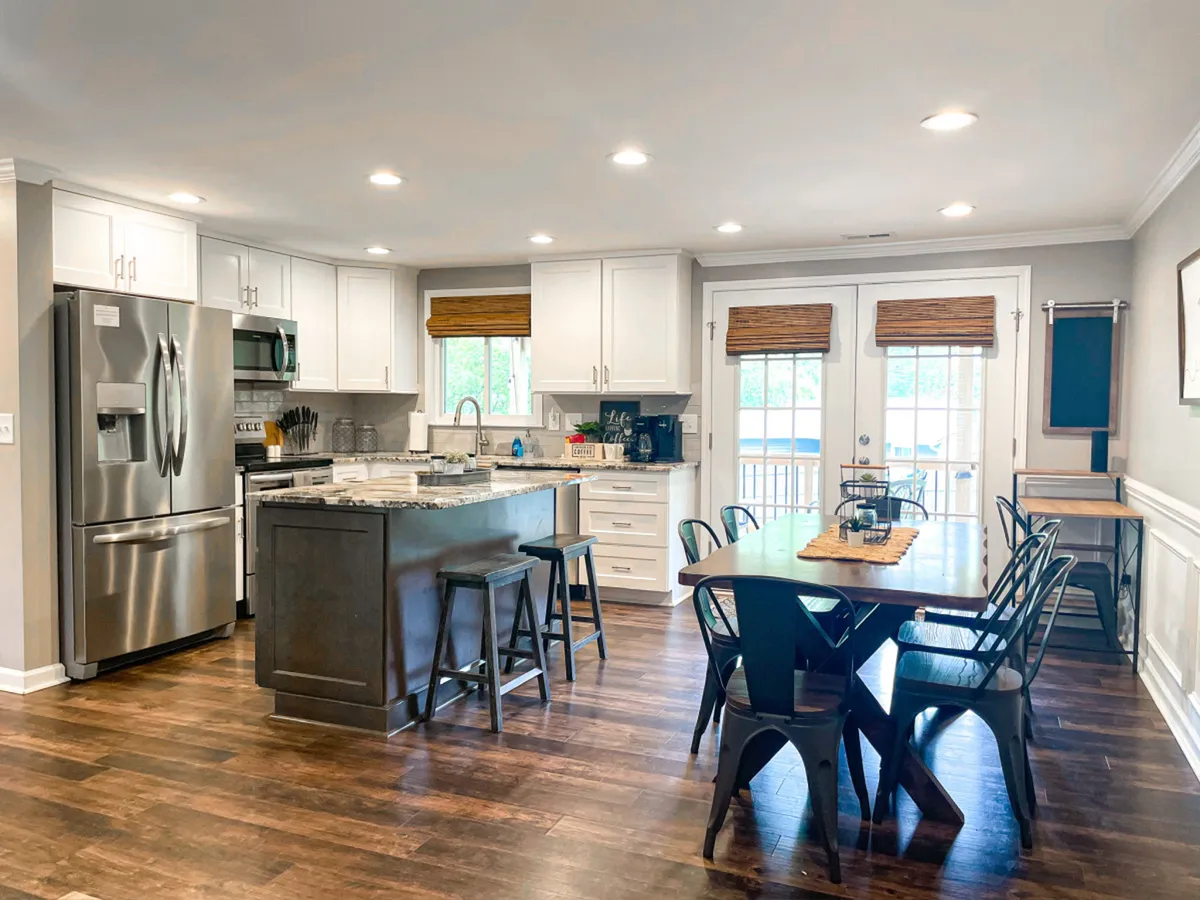 A warm and welcoming view featuring a kitchen island and a spacious dining table, with stainless steel appliances gleaming in the background and a window letting in natural light. The open layout visually connects the kitchen with the adjoining dining area, inviting both cooking and social interaction.