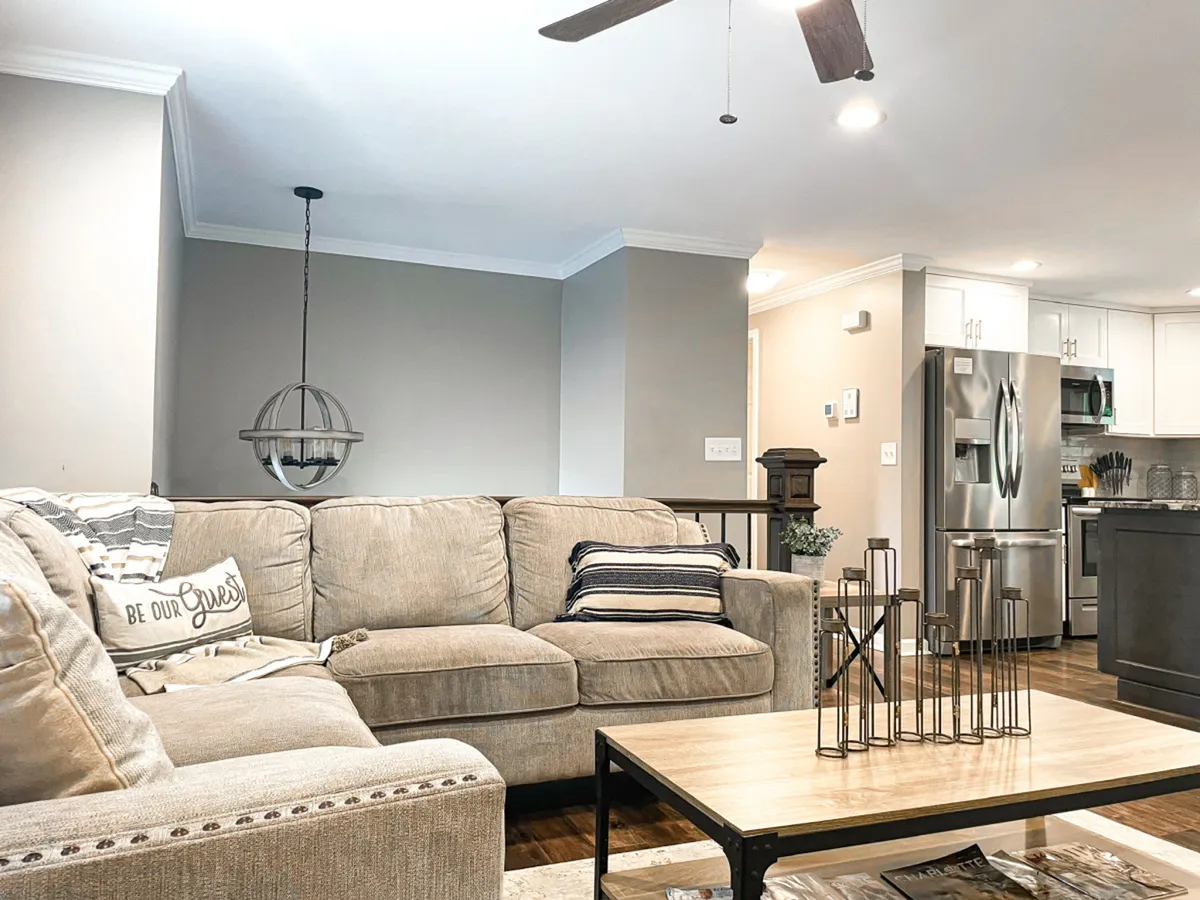A bright and welcoming living space is depicted, complete with a comfortable sofa, stylish lighting, modern kitchen appliances, and a coffee table arranged with decorative items. The scene suggests a space perfect for relaxing, entertaining, or sharing a meal.