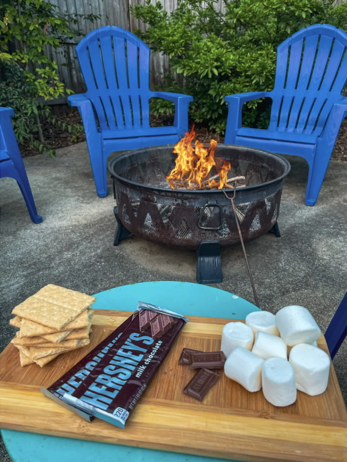 A central fire pit surrounded by blue chairs, with s’mores ingredients placed nearby, set against a backdrop of lush greenery. The setup invites guests to gather for cozy evenings under the stars, enjoying warmth and conversation.