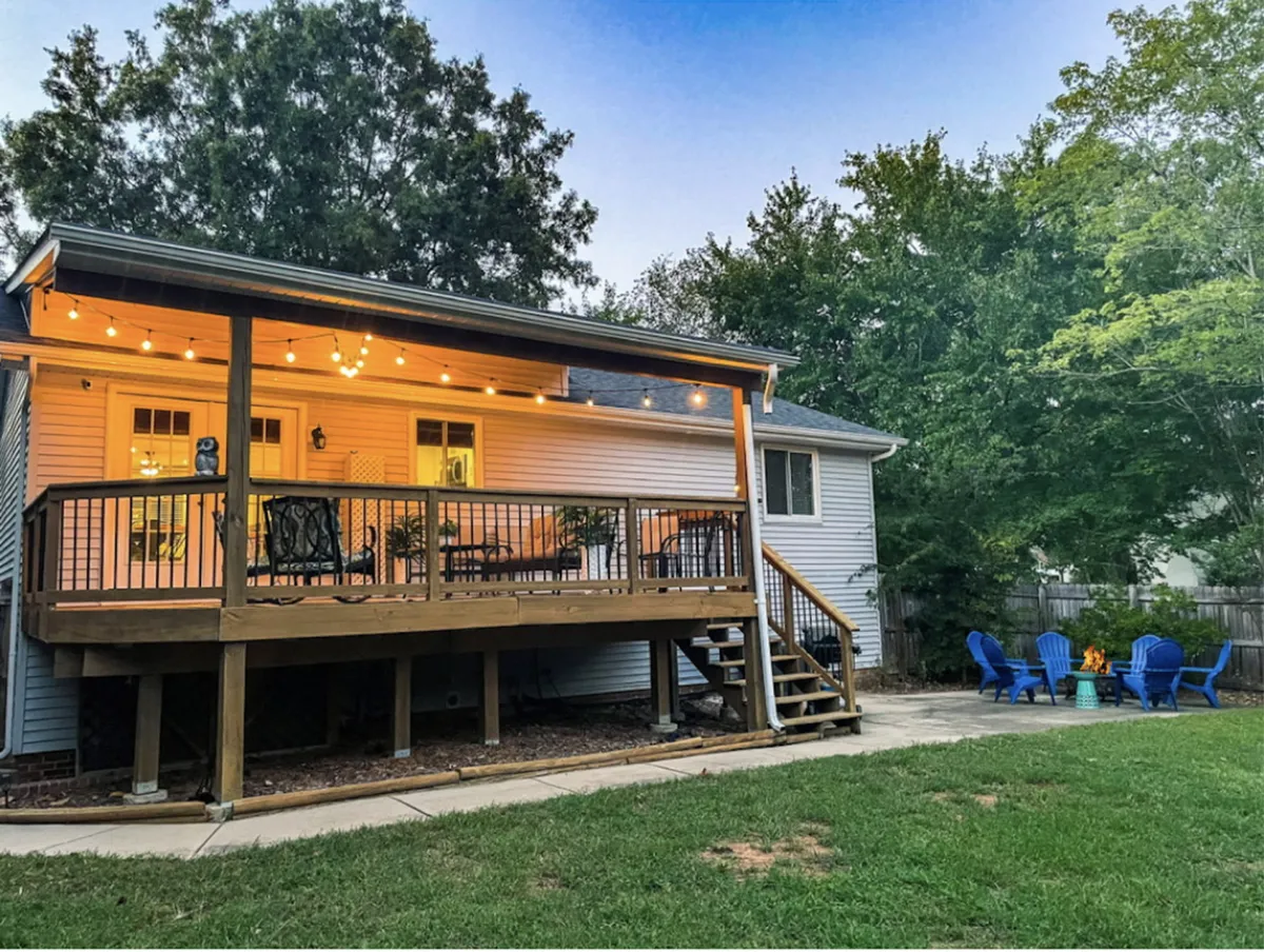 An exterior view of a deck adorned with decorative string lights, blue Adirondack chairs, and a fire pit. The scene captures the charm of evening outdoor gatherings and the warmth of shared moments by the fire.