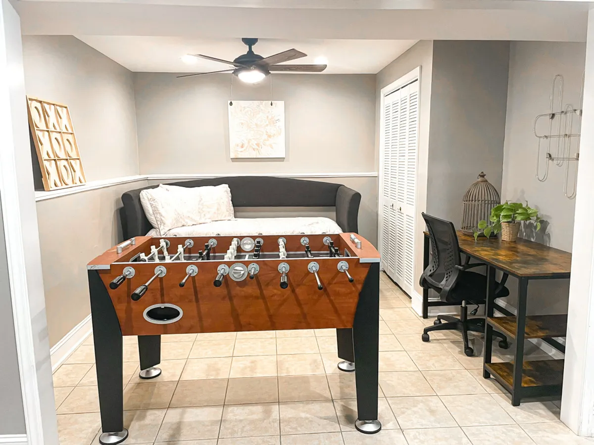 A fun and casual interior space featuring a foosball table, a comfortable sectional sofa, and a workspace desk off to the side. The room is set up for both friendly competition and relaxed downtime with friends or colleagues.
