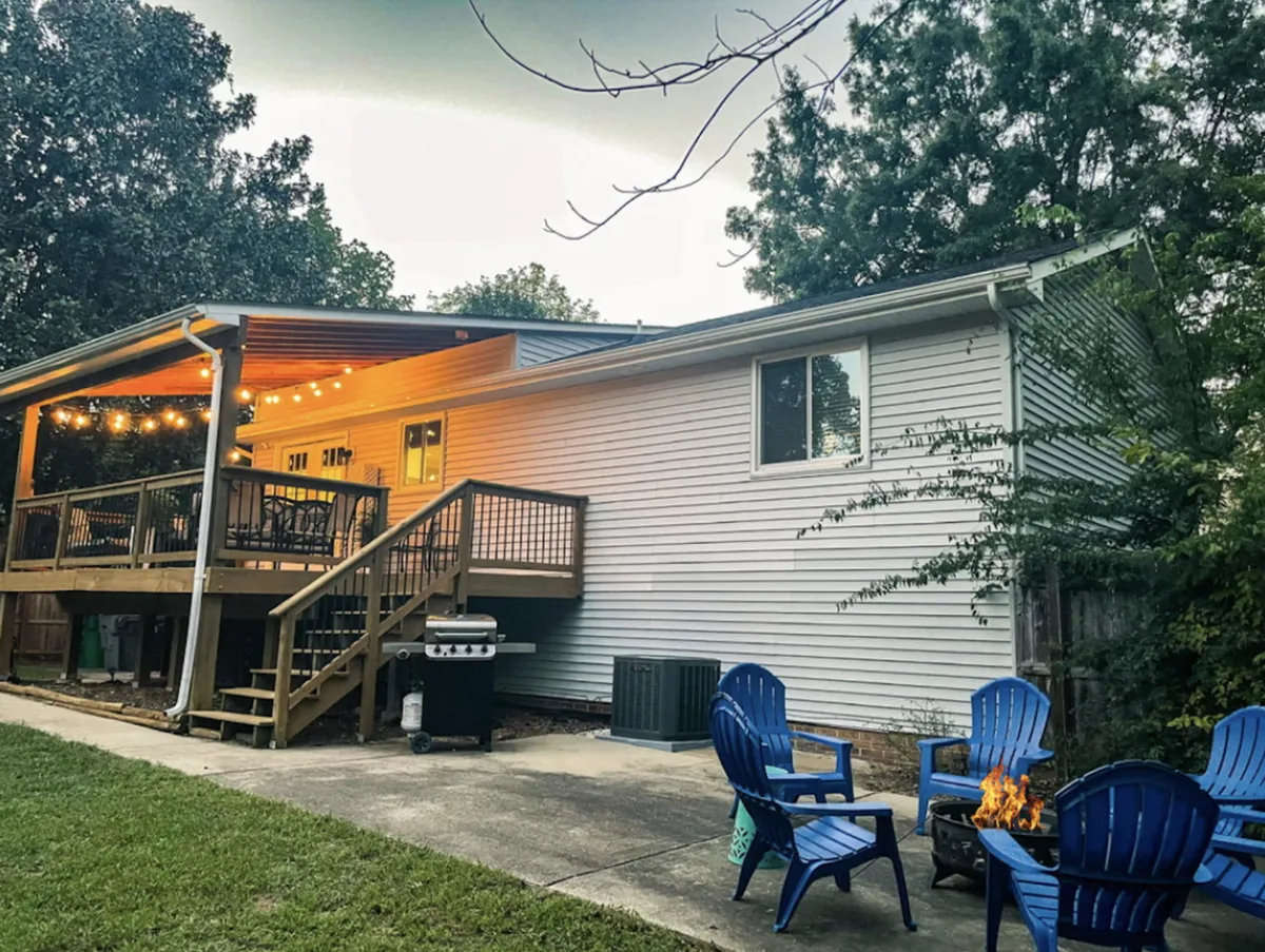 This exterior image features a spacious deck adorned with string lights, inviting blue Adirondack chairs, and a central fire pit. The setup conveys a relaxed, communal atmosphere perfect for evening gatherings, socializing, or unwinding after a day of adventure. The lush backdrop suggests a private backyard setting ideal for family activities and outdoor fun.