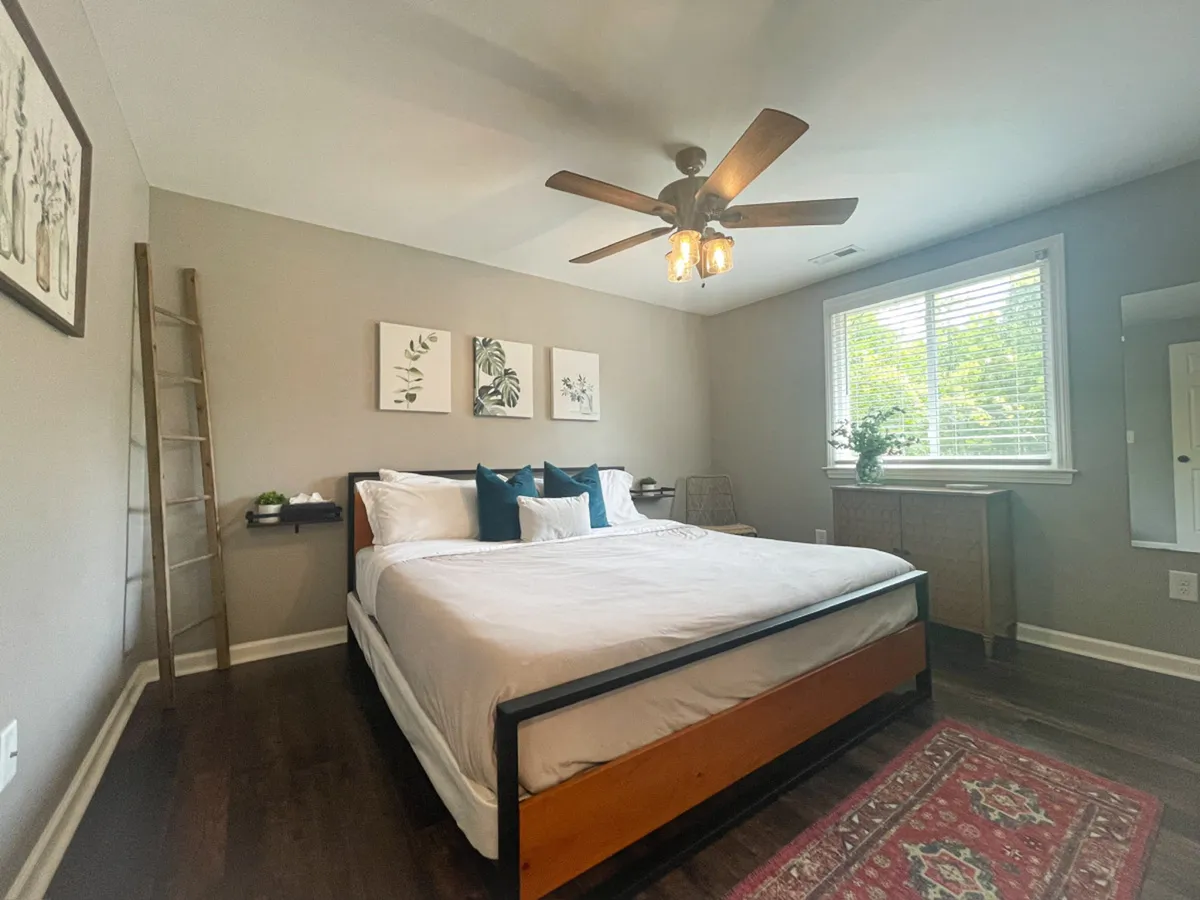 A spacious bedroom featuring a large bed with crisp white bedding, blue accent pillows, wall art, a window streaming natural light, and a wooden dresser. The atmosphere is tranquil and inviting, suggesting a restful retreat.