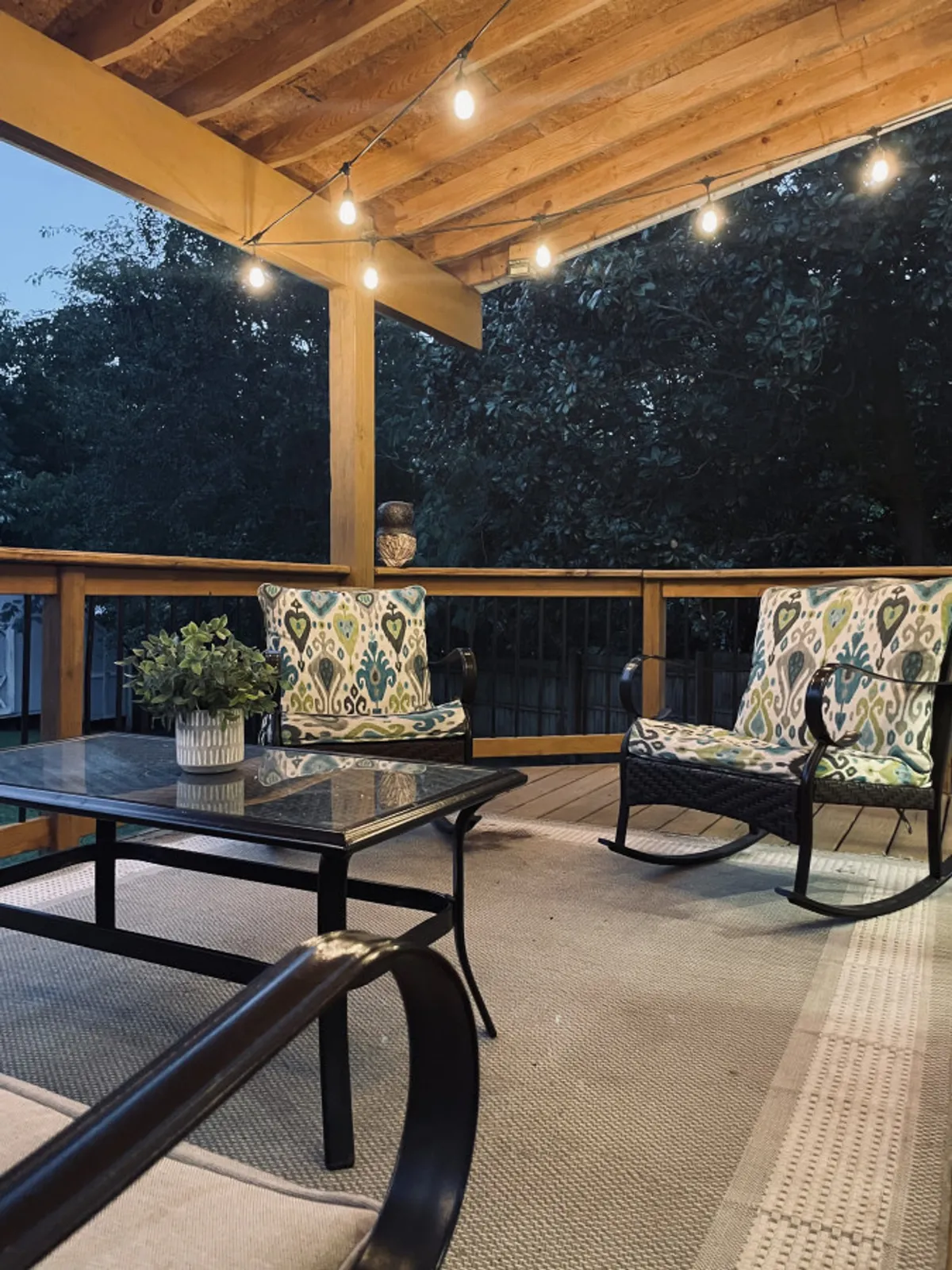 An inviting exterior covered patio featuring string lights overhead, rocking chairs positioned for conversation, and a decorative plant at one end. The space exudes warmth and is clearly designed for evening gatherings and relaxed entertainment.