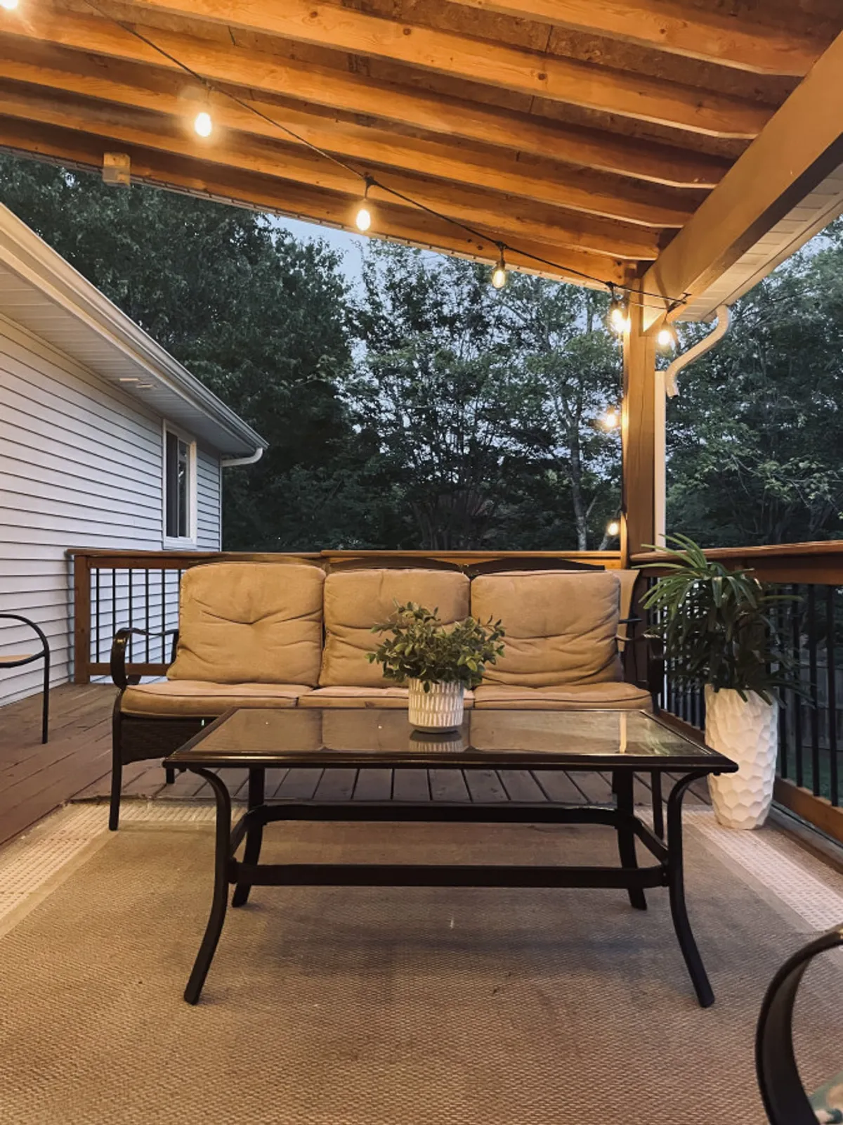 A cozy outdoor seating area with a sofa, coffee table, potted plant, and string lights overhead. The setup invites guests to socialize, relax, or share stories in an inviting exterior environment.