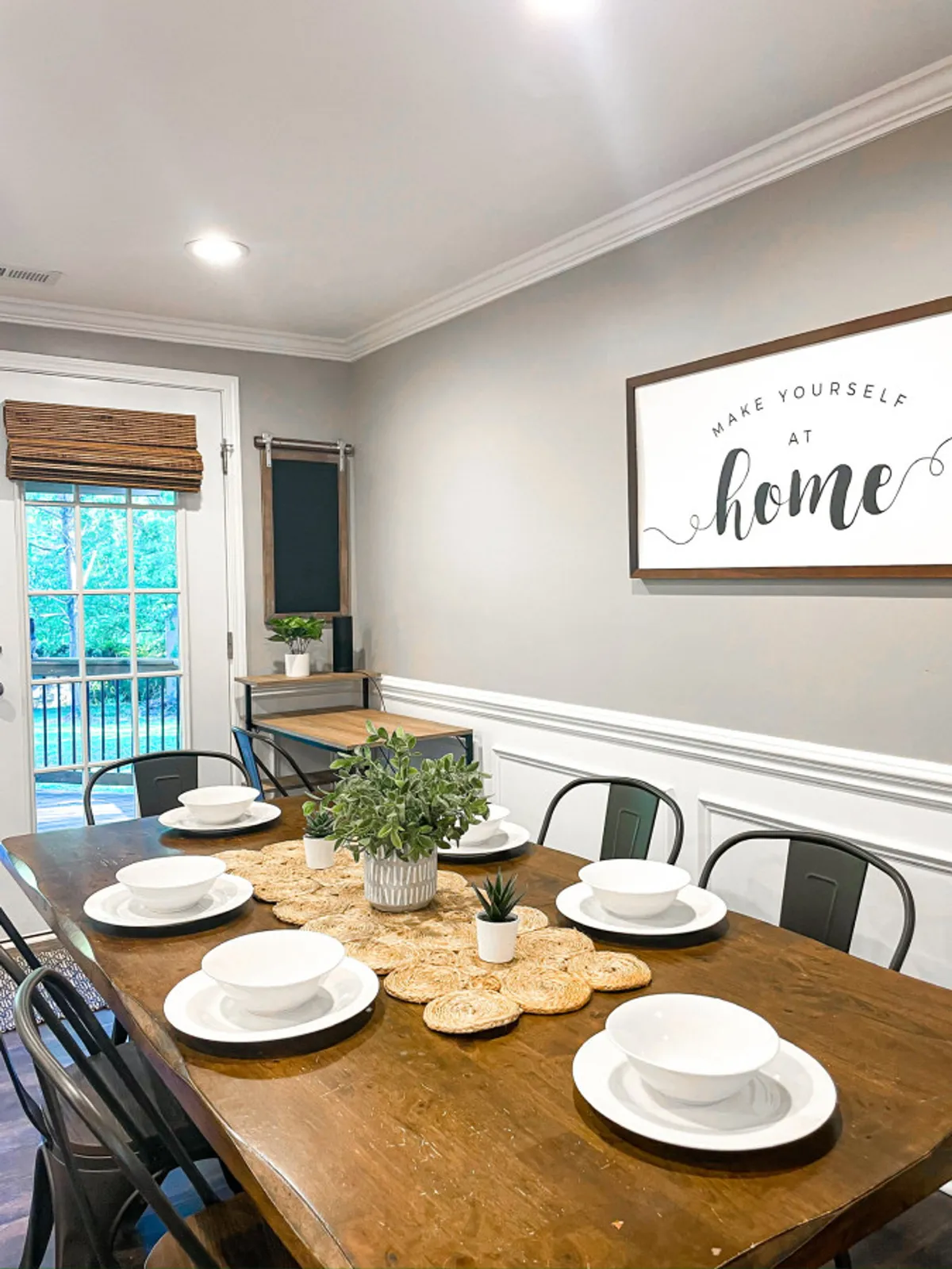 A cozy dining area centered around a table set for a meal, accentuated by plants and a decorative sign, exuding a sense of warmth and togetherness. The inviting place settings evoke the idea of shared meals and casual gatherings.