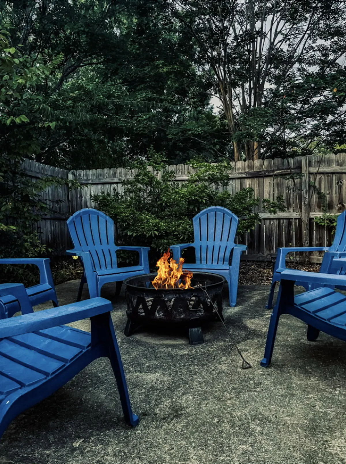 A tranquil backyard scene with a campfire surrounded by blue chairs set amidst lush greenery. The campfire is the focal point, evoking images of storytelling, connection, and enjoying the natural surroundings together as a family or group.