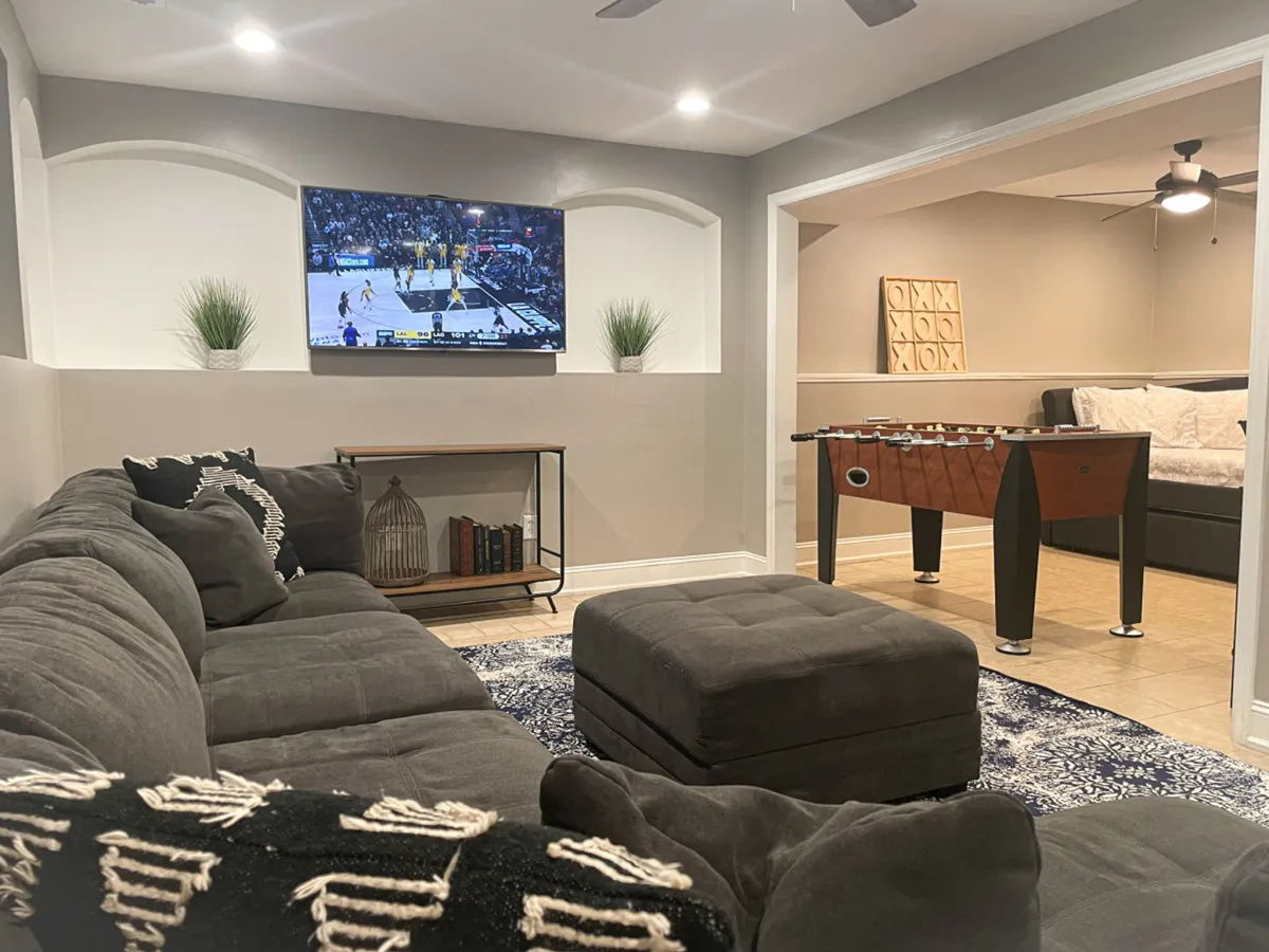This image features a large sectional couch arranged around a flat-screen TV, with a foosball table nearby and decorative plants adding a touch of warmth. The scene is casual and friendly, suggesting an inviting atmosphere for relaxation, watching TV, or enjoying games with friends and family.